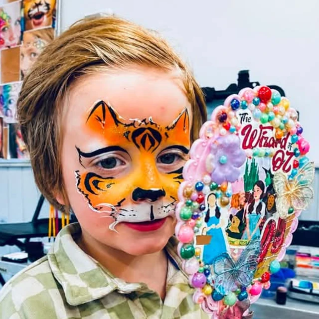A child with orange tiger face paint holding a colourful Wizard of Oz mirror