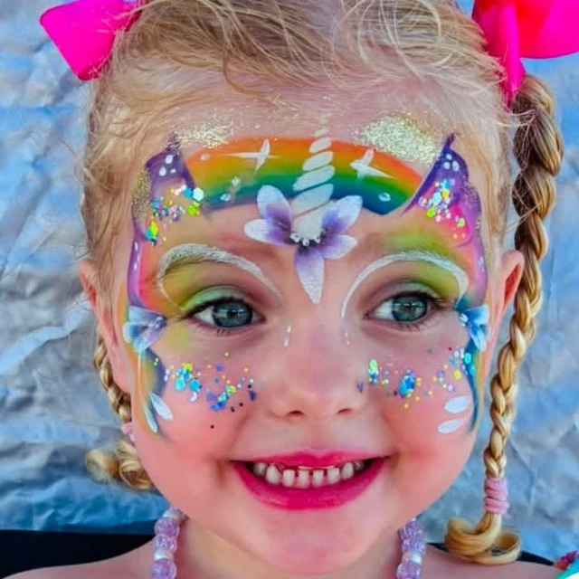 A smiling child with rainbow unicorn face paint and sparkling flowers